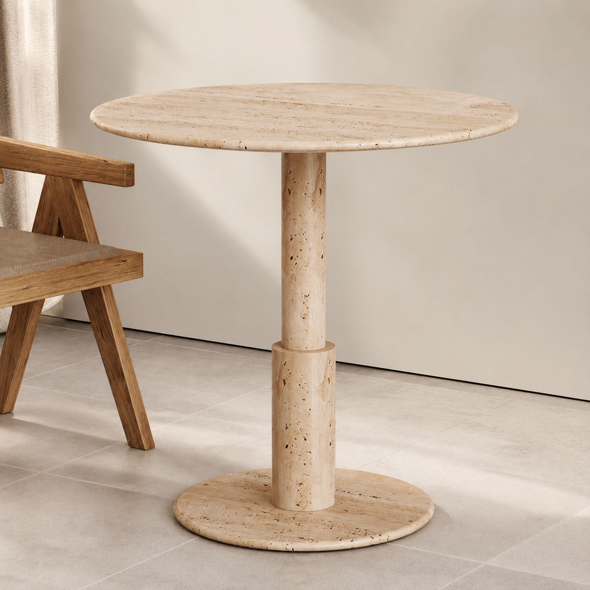 RoundTwin Elite beige travertine pedestal dining table beside a wooden armchair in a neutral interior.