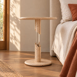 AeroCloud travertine side table with a transparent acrylic support detail beside a linen bed, shown from a low front view.