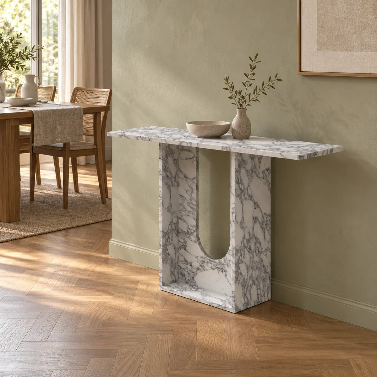 Right-front view of the Alecto Arabescato marble console beside a warm dining room, showing its thin square-ended top, deep stone side and rounded open-centre base.
