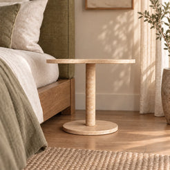Low frontal view of the CloudDream travertine side table beside a linen bed.
