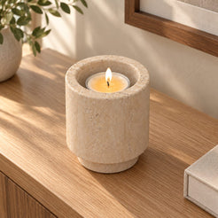 Tall RoundHaven Classico travertine candle holder with one lit tealight, high left oblique view in a warm sunlit interior