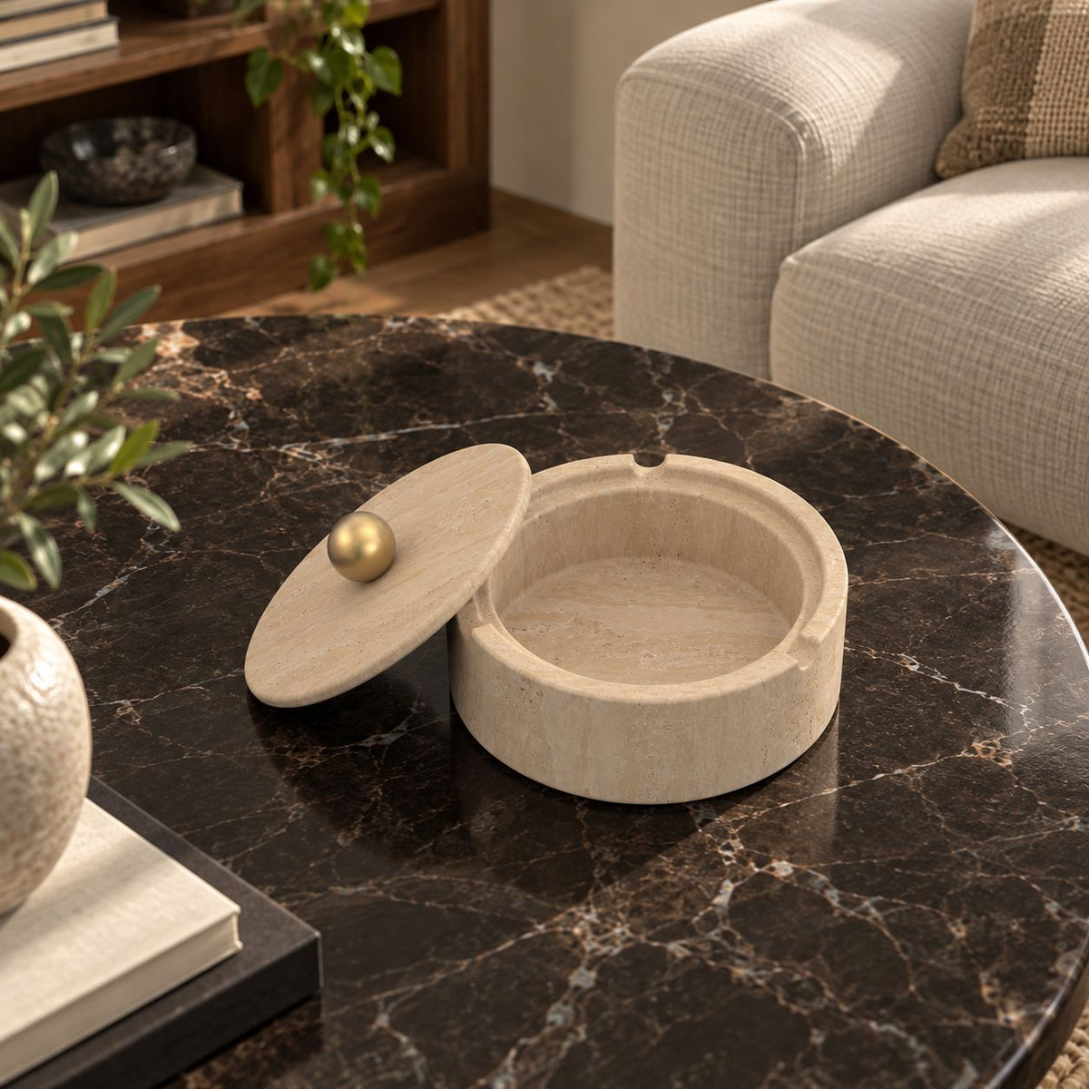 RoundHaven Classico travertine ashtray with one matching gold-knob lid, high left oblique view in a warm natural-light interior