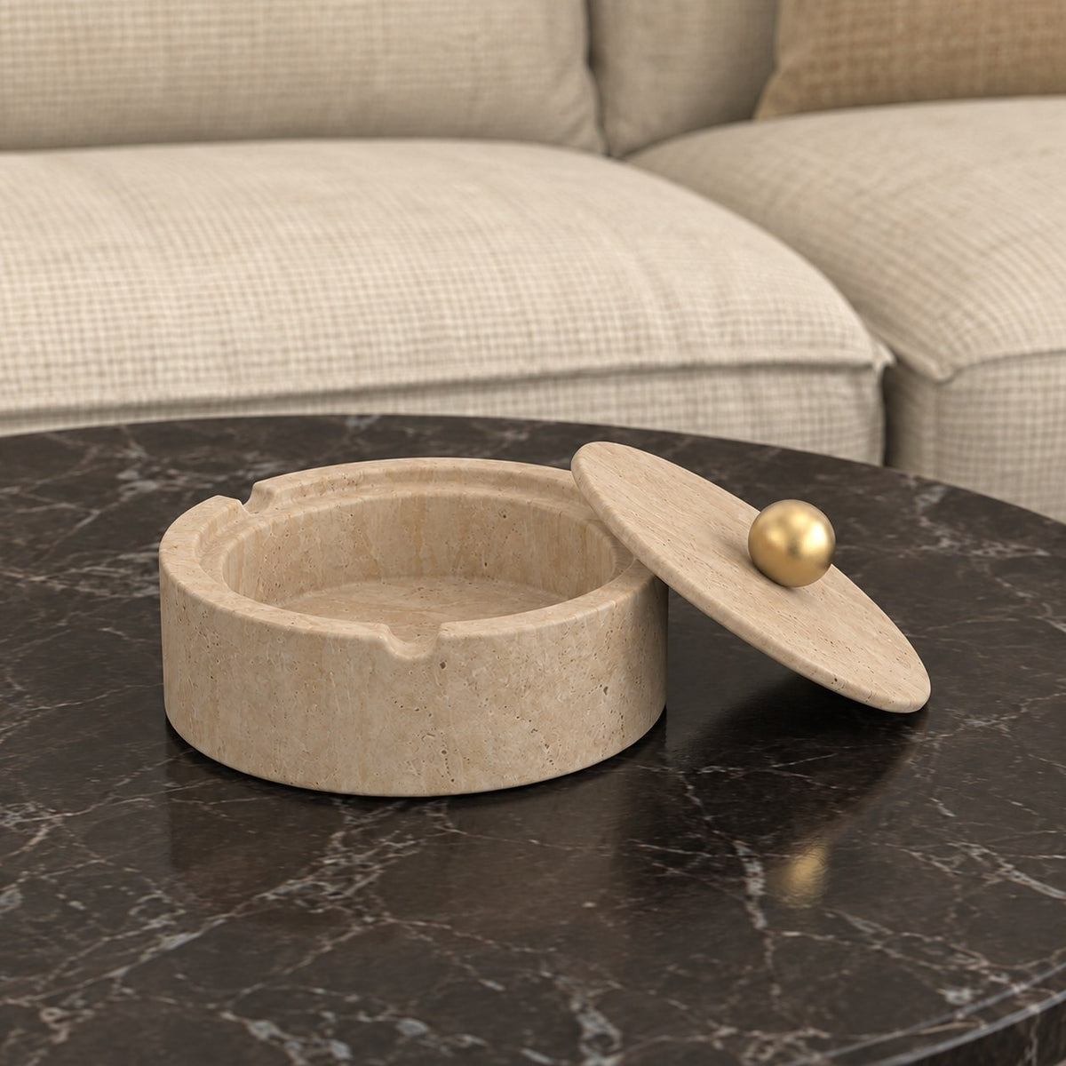 RoundHaven Classico travertine ashtray and matching gold-knob lid, viewed from the right on a dark stone coffee table