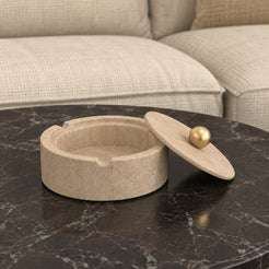 RoundHaven Classico travertine ashtray and matching gold-knob lid, viewed from the right on a dark stone coffee table