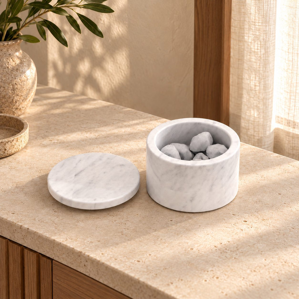 RoundHaven Carrara marble aroma diffuser with its matching flat lid and source styling stones, left oblique view in a warm sunlit interior