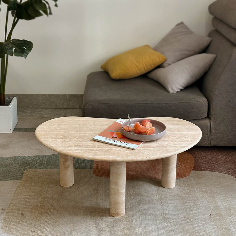 PeaPod Oval Travertine Coffee Table placed beside a cozy sofa, adorned with a magazine and fruit bowl, highlighting its functional and aesthetic appeal.