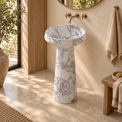 Pala Italian Arabescato Marble Pedestal Sink with a shallow round basin and tapered cylindrical pedestal in a warm cream and natural oak bathroom, three-quarter view.
