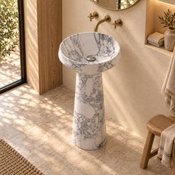 Pala Italian Arabescato Marble Pedestal Sink in a warm cream and oak bathroom, elevated view showing its round basin, silver drain cover and full tapered stone pedestal.