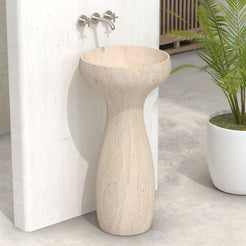 Angled view of the Pava travertine pedestal sink beside a potted palm