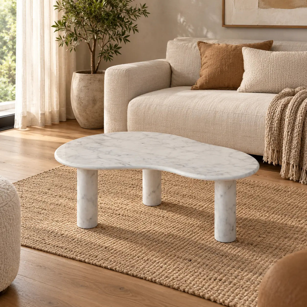 PeaPod Italian Carrara marble coffee table with a thin curved top and three cylindrical stone legs in a warm linen-sofa living room.