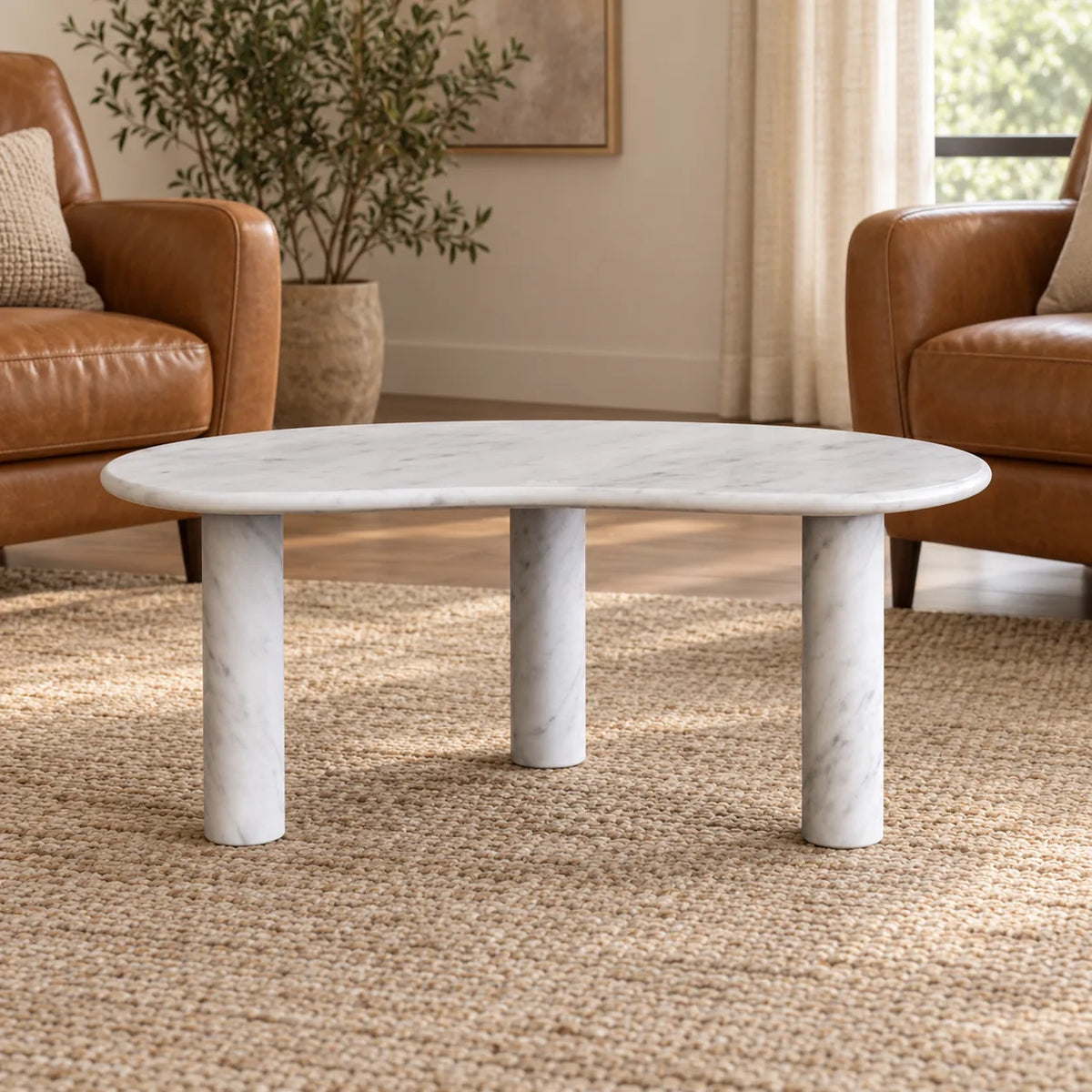 Low front view of the PeaPod Carrara marble coffee table in a leather-chair reading area, showing its thin bean-shaped top and three stone supports.
