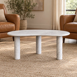 Low front view of the PeaPod Carrara marble coffee table in a leather-chair reading area, showing its thin bean-shaped top and three stone supports.