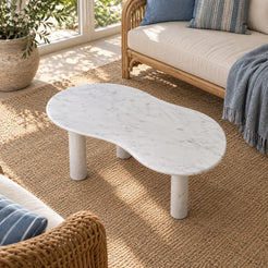 Elevated view of a single PeaPod Carrara marble coffee table in a garden-facing sunroom, showing its curved stone top and three cylindrical legs.