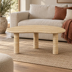 Low front view of the PeaPod Classico travertine coffee table in a linen-sofa living room, showing its thin bean-shaped top and three stone supports.