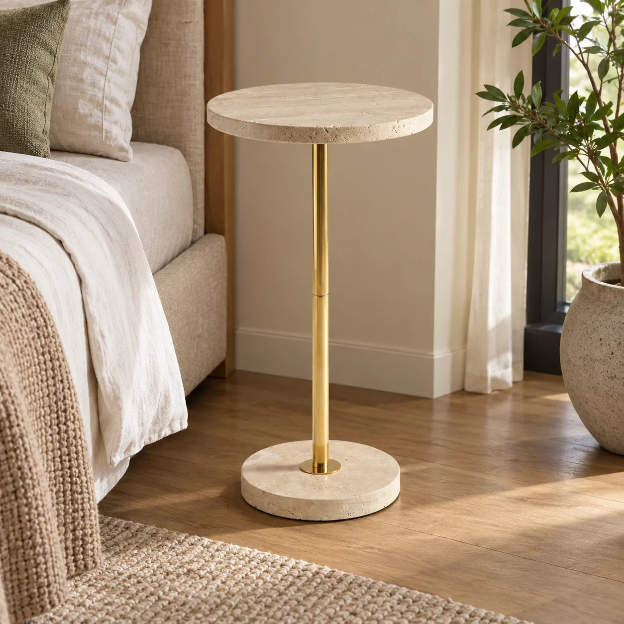 Pillar side table with a Classico travertine top and gold-finish metal stand beside a linen bed.