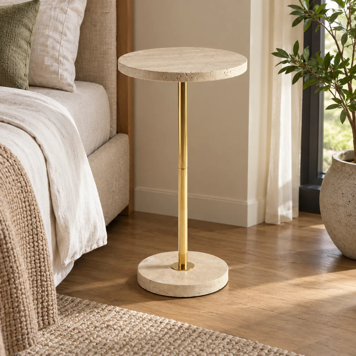 Pillar side table with a Classico travertine top and gold-finish metal stand beside a linen bed.