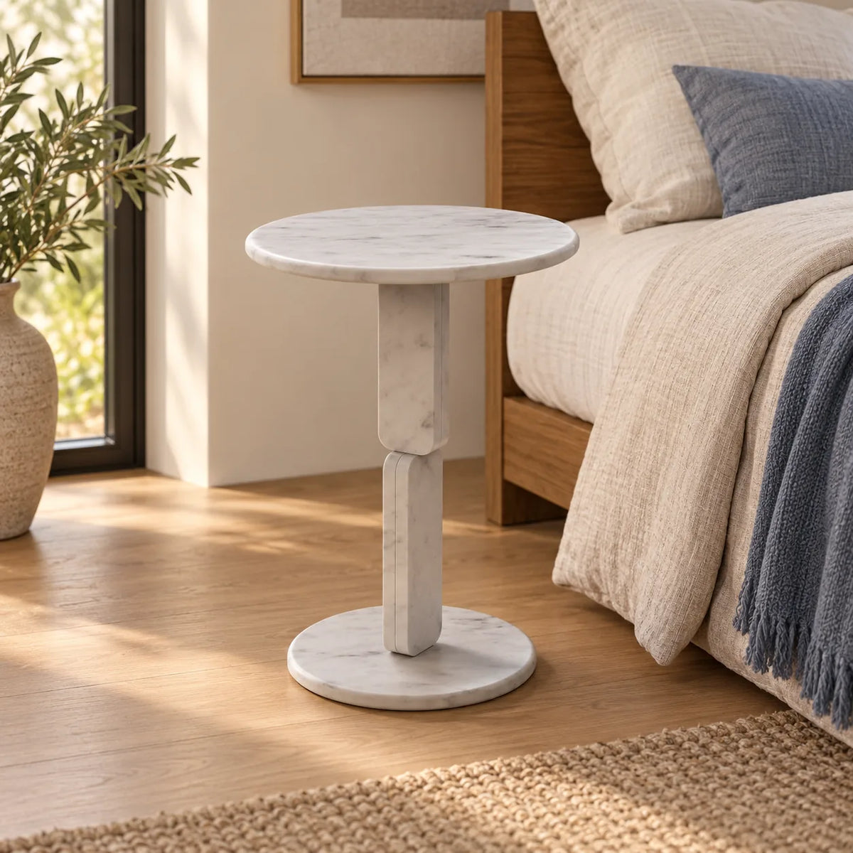 RoundGeo Carrara marble side table beside a linen bed, shown from a moderately elevated left-front view