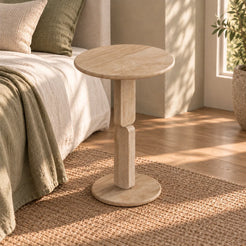 RoundGeo Italian Classico travertine side table beside a linen bed, shown from a moderate front-quarter view
