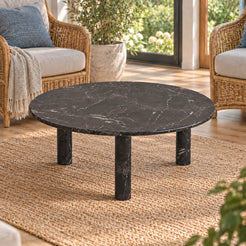 Elevated view of the RoundHaven Spanish Dark Emperador marble coffee table in a garden-view sunroom, showing its circular stone top and three cylindrical supports.