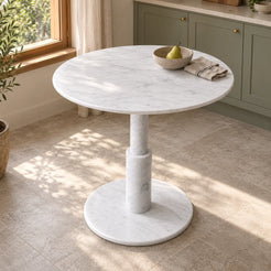 RoundTwin Elite Carrara white marble dining table from an elevated oblique angle in a sage and oak kitchen breakfast corner, showing the round top and stepped cylindrical pedestal