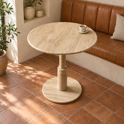 RoundTwin Elite beige travertine bistro dining table, high oblique view showing the stepped cylindrical stem and round floor base in a warm cafe alcove