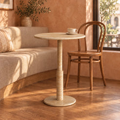 RoundTwin Small Travertine Dining Table in a warm cafe breakfast nook, low view showing the thin round stone top, slender stepped stem and circular floor base
