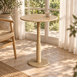 RoundTwin Small Travertine Dining Table beside a garden-facing sunroom window, moderately elevated view of the round beige stone top, slender stepped stem and circular base