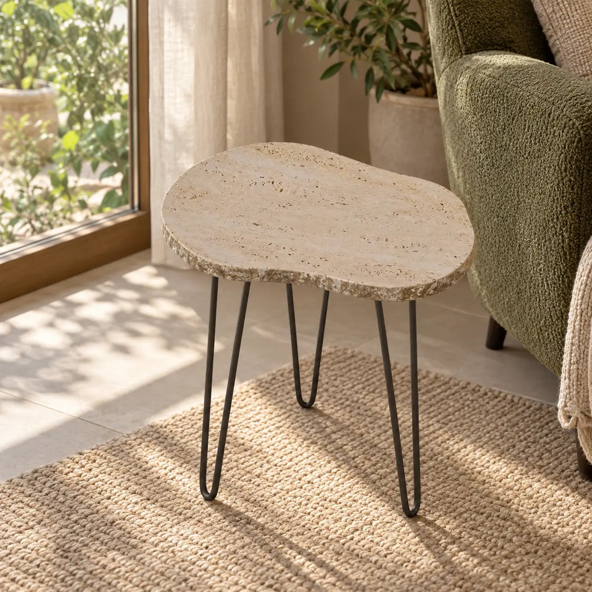 StoneMist travertine side table beside a moss reading chair, shown from a high front-quarter view with its irregular tabletop and hairpin supports