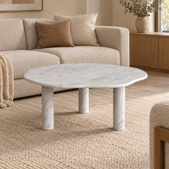 SunAura Carrara marble coffee table with a softly lobed top and three cylindrical legs in a warm living room.