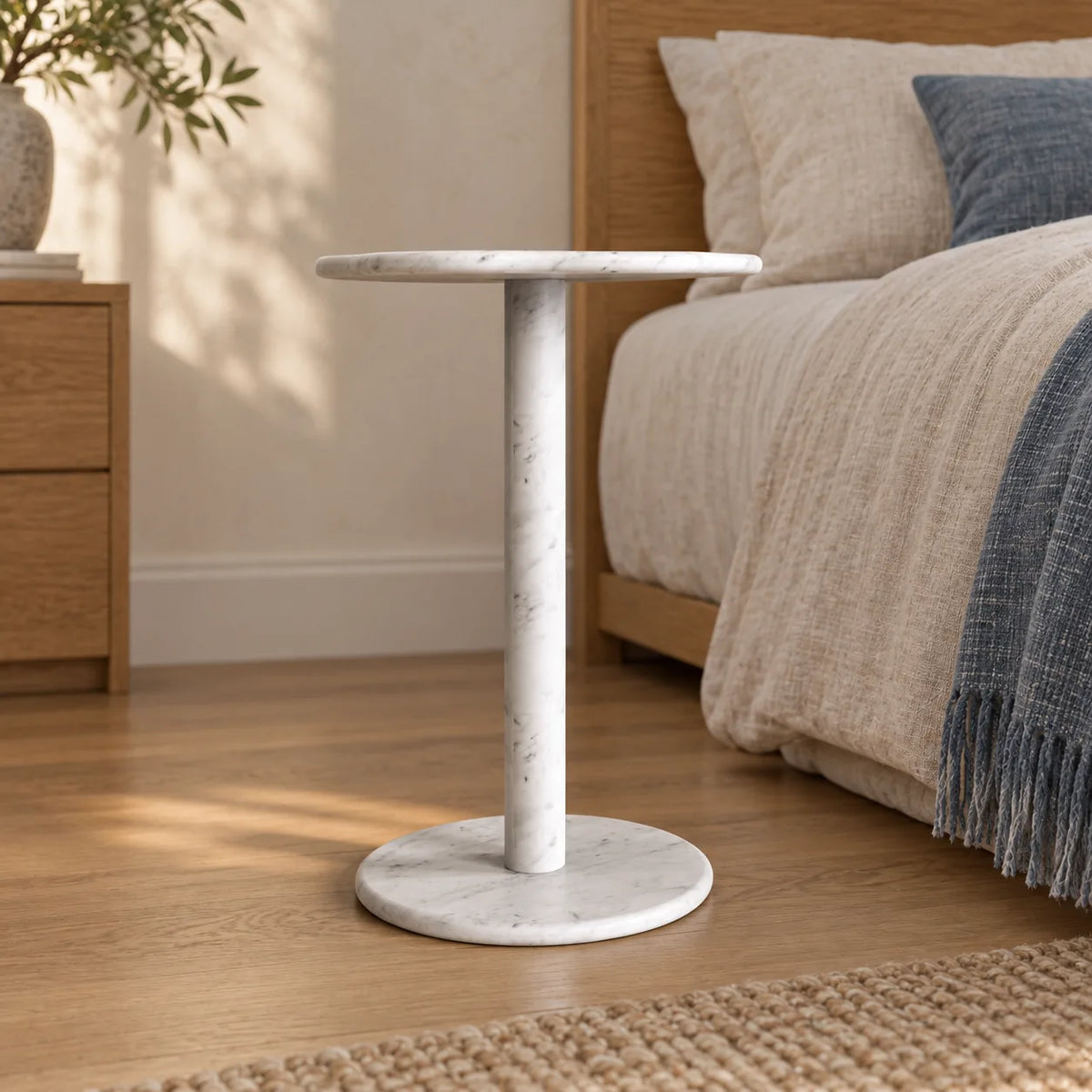 SunAura Carrara marble side table beside a linen bed, shown from a low front view