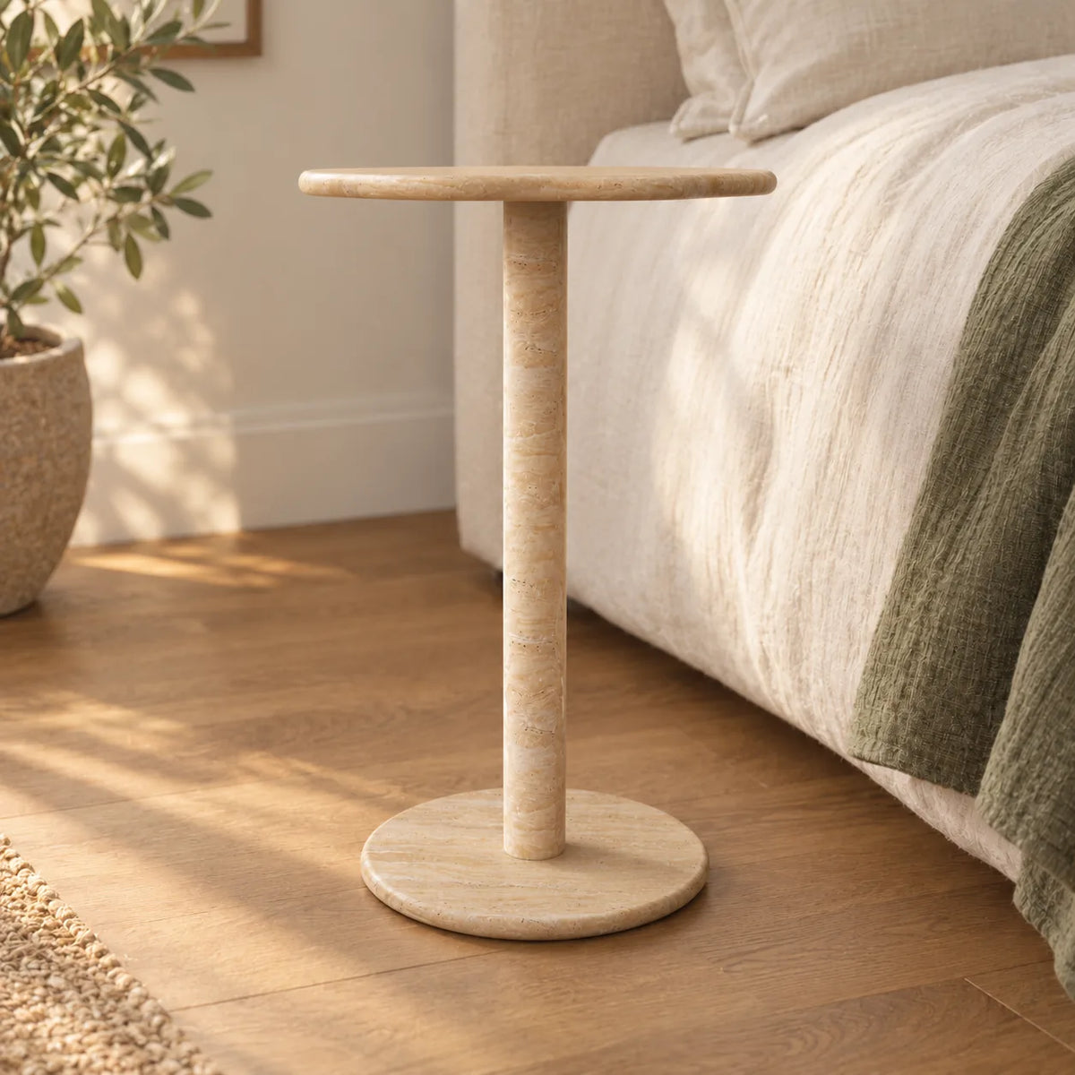 SunAura Italian Classico travertine Standard side table beside a linen bed, shown from a low front view