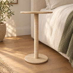 SunAura Italian Classico travertine Standard side table beside a linen bed, shown from a low front view