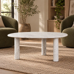Low front view of the SwanEgg Carrara marble coffee table in a green-chair reading nook, showing its thin egg-shaped top and three round stone supports.