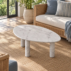 Elevated view of the SwanEgg Carrara marble coffee table in a garden-facing sunroom, showing the soft egg-shaped top and three cylindrical legs.
