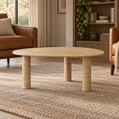 Low front view of the SwanEgg Classico travertine coffee table in a leather-chair reading area, showing its thin egg-shaped top and three stone supports.