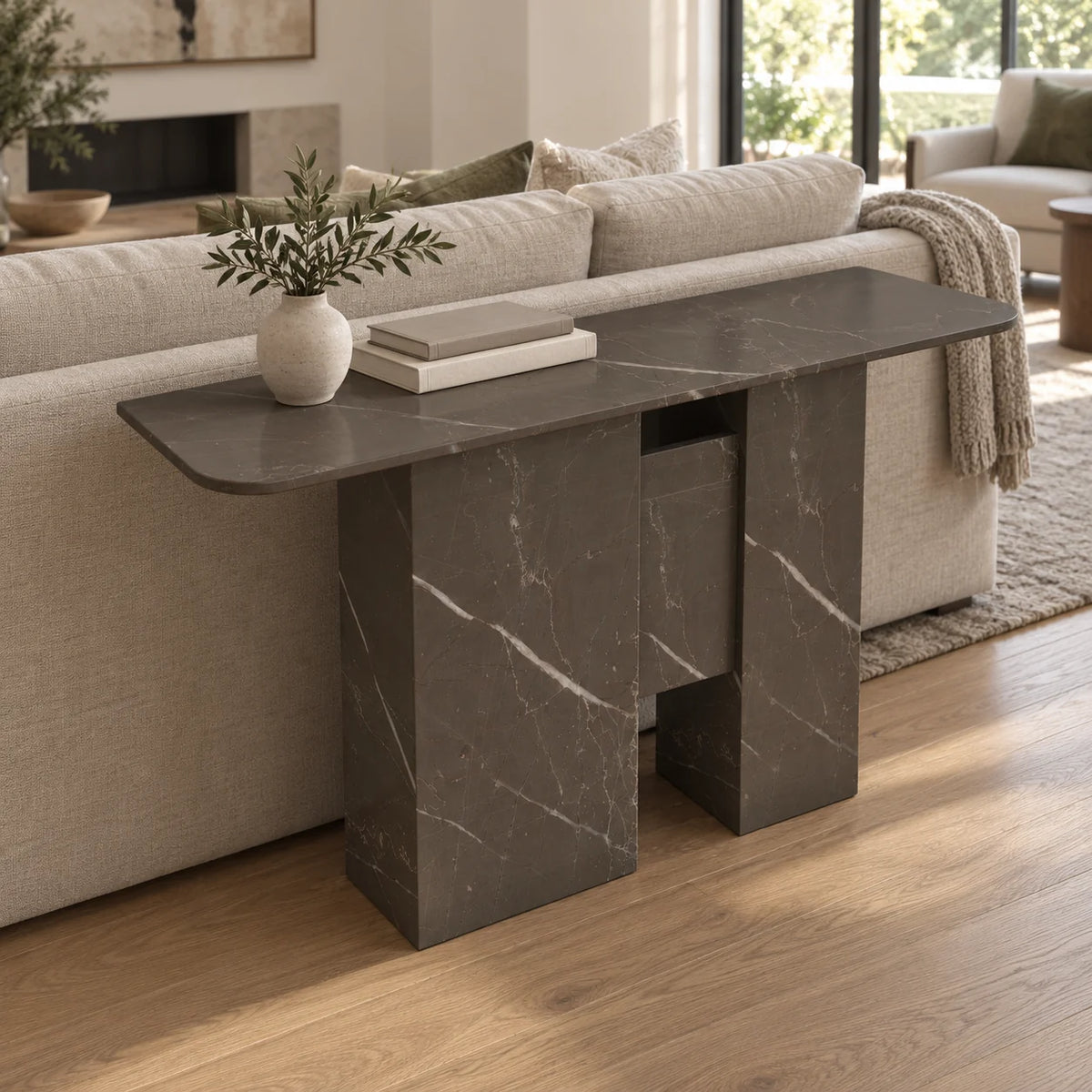 Elevated left-front view of the Thalia Bulgarian Gray marble console behind a linen sofa, showing its rounded-end top, stone supports and suspended central panel