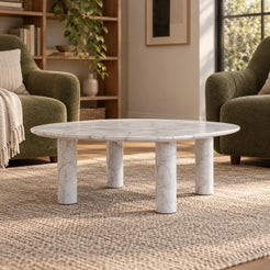 Low front view of the ZenPebble Carrara marble coffee table in a green-chair reading lounge, showing its rounded-square top and four stone supports.