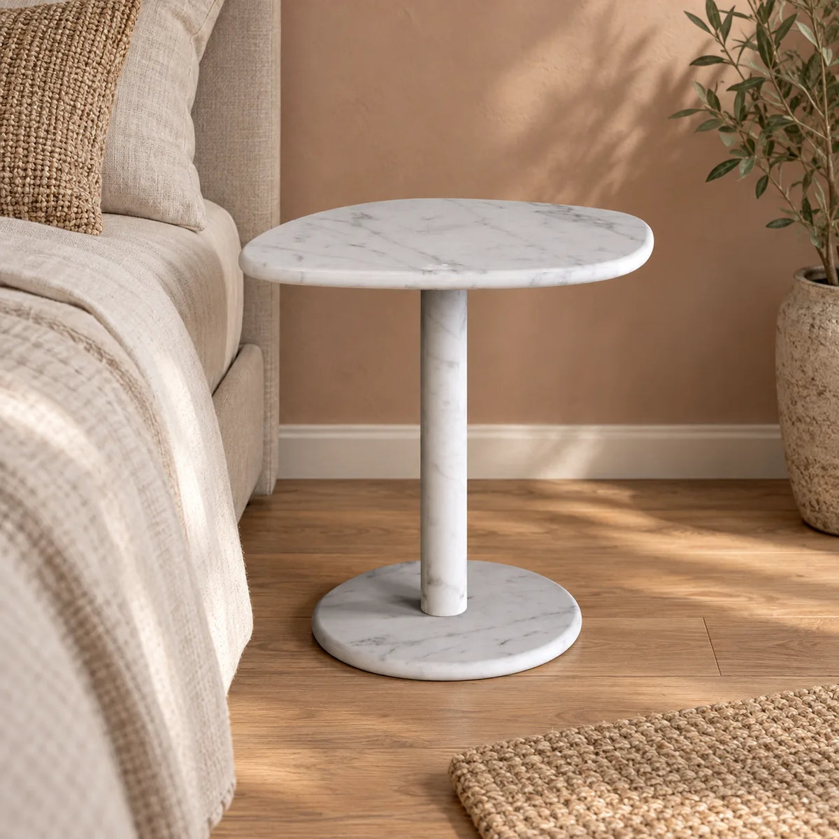 ZenPebble Carrara marble side table beside a bed, shown from a low frontal view with the full pedestal and base visible