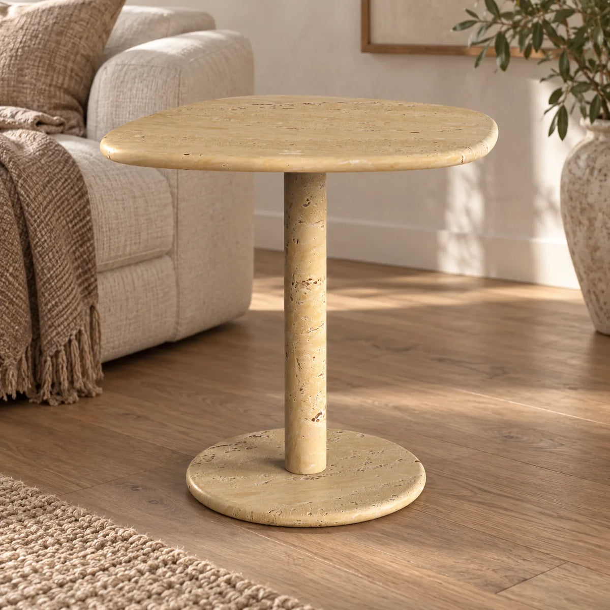 ZenPebble Classico travertine side table with a softly rounded square top and circular base beside a linen sofa.