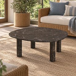 Moderate oblique view of the ZenPebble Dark Emperador marble coffee table in a garden-facing sunroom, showing its soft-square top and cylindrical stone supports.